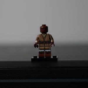 May include: A small plastic minifigure with a dark brown head and torso, wearing a tan tunic with a wrap-style neckline and a dark brown belt. The figure has dark brown legs with white accents at the knees and stands on a black rectangular base.