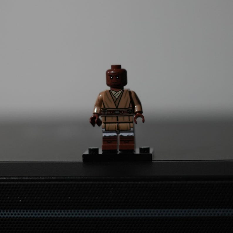 May include: A small plastic minifigure with a dark brown head and torso, wearing a tan tunic with a wrap-style neckline and a dark brown belt. The figure has dark brown legs with white accents at the knees and stands on a black rectangular base.