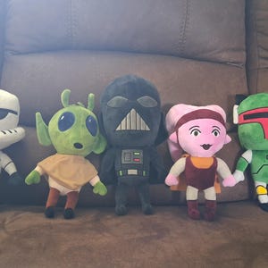 Pode incluir: Um conjunto de cinco peluches representando personagens de Star Wars. A coleção inclui um stormtrooper branco, um alienígena verde com olhos grandes, uma figura preta de Darth Vader, uma personagem Twi'lek rosa e um caçador de recompensas Boba Fett verde e vermelho.