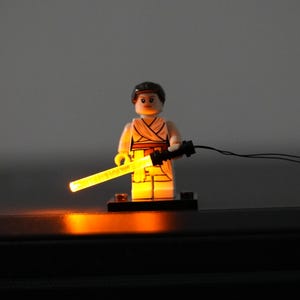 May include: A plastic minifigure with dark brown hair, wearing a tan outfit with a wrap-style top, standing on a black base. The figure holds a black handle with a glowing, translucent yellow light-up rod extending from it, connected by a thin wire.