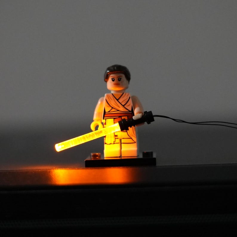 May include: A plastic minifigure with dark brown hair, wearing a tan outfit with a wrap-style top, standing on a black base. The figure holds a black handle with a glowing, translucent yellow light-up rod extending from it, connected by a thin wire.