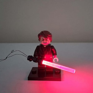 May include: A plastic minifigure with brown hair and a stern facial expression, dressed in a black outfit. The figure holds a glowing red lightsaber accessory that emits a bright light, standing on a small black rectangular display base.
