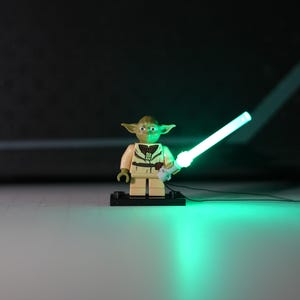 May include: A small green plastic character figurine with pointed ears, wearing a tan tunic with brown details, standing on a black rectangular base. The figure holds a bright, glowing green light-up sword that illuminates the surface below.