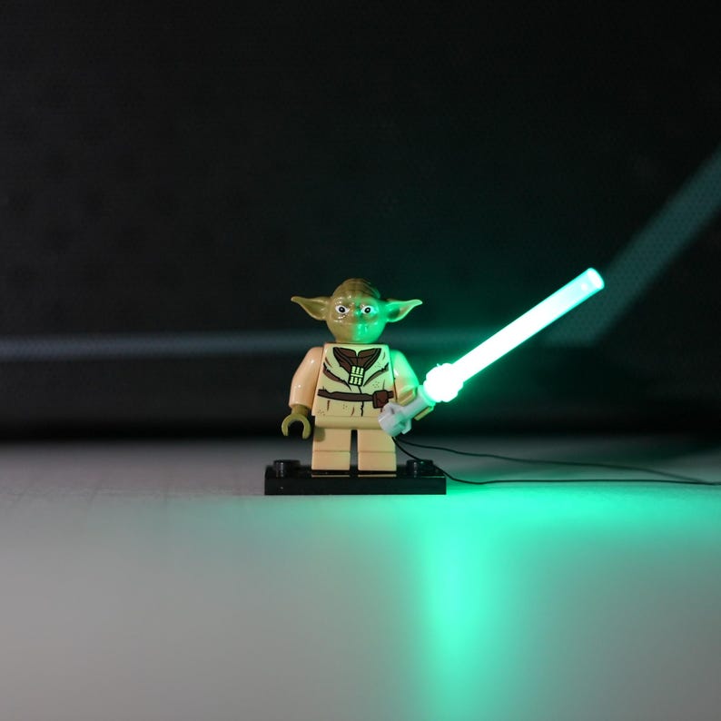 May include: A small green plastic character figurine with pointed ears, wearing a tan tunic with brown details, standing on a black rectangular base. The figure holds a bright, glowing green light-up sword that illuminates the surface below.
