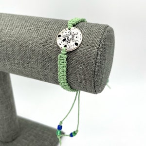 May include: A green braided bracelet with a silver sand dollar charm and blue and green beads.