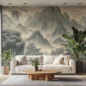 以下が含まれることがあります： クリーム色のソファが、緑の木々と枝垂れ柳が描かれた霧深い山々の壁画の前に置かれています。ソファの前には円形の木製コーヒーテーブルが中央に配置され、その両脇に2つの緑の鉢植えが置かれています。