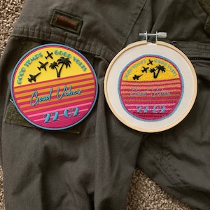 May include: Two embroidered patches featuring a sunset and palm trees design. The patches are on a green fabric background. The text on the patches reads "Good Vibes" and "22-02".