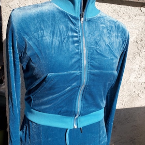 May include: A teal velour tracksuit, featuring a zip-up jacket with a high collar and a matching pair of pants. The jacket has a front pocket and a silver zipper. The outfit is displayed on a mannequin, showcasing the soft texture and color.