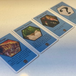 Puede incluir: Cuatro cartas de juego azules con diferentes im&aacute;genes y texto. Las cartas son para un juego de mesa llamado "King's Wild". Las cartas est&aacute;n dispuestas en fila sobre una superficie blanca. El texto en las cartas dice: "Pillage", "Dominio Eminente", "King's Wild" y "?".