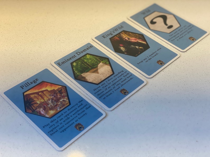 Puede incluir: Cuatro cartas de juego azules con diferentes im&aacute;genes y texto. Las cartas son para un juego de mesa llamado "King's Wild". Las cartas est&aacute;n dispuestas en fila sobre una superficie blanca. El texto en las cartas dice: "Pillage", "Dominio Eminente", "King's Wild" y "?".