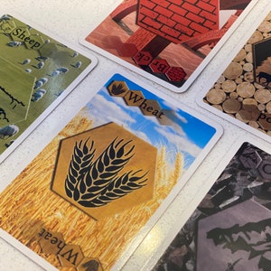 Puede incluir: Cinco cartas de juego diferentes con im&aacute;genes de ovejas, trigo, ladrillos, madera y mineral. Las cartas son del juego de mesa Los Colonos de Cat&aacute;n.