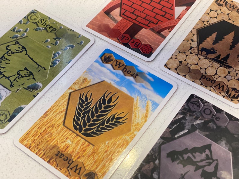 Puede incluir: Cinco cartas de juego diferentes con im&aacute;genes de ovejas, trigo, ladrillos, madera y mineral. Las cartas son del juego de mesa Los Colonos de Cat&aacute;n.