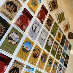 Puede incluir: Una colecci&oacute;n de cartas coloridas con varias im&aacute;genes y texto. Las cartas presentan im&aacute;genes de edificios, recursos y otros elementos del juego. El texto en las cartas incluye los nombres de las cartas, as&iacute; como las reglas e instrucciones del juego. Las cartas est&aacute;n dispuestas en un patr&oacute;n de cuadr&iacute;cula sobre una superficie blanca.