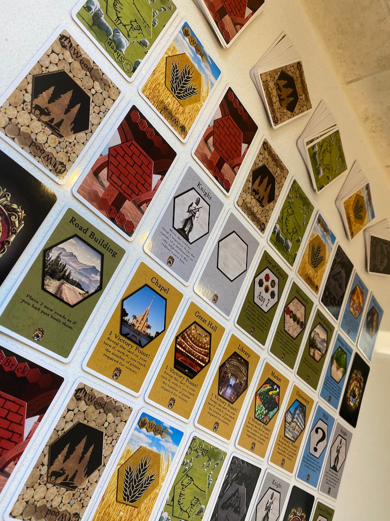 Puede incluir: Una colecci&oacute;n de cartas coloridas con varias im&aacute;genes y texto. Las cartas presentan im&aacute;genes de edificios, recursos y otros elementos del juego. El texto en las cartas incluye los nombres de las cartas, as&iacute; como las reglas e instrucciones del juego. Las cartas est&aacute;n dispuestas en un patr&oacute;n de cuadr&iacute;cula sobre una superficie blanca.