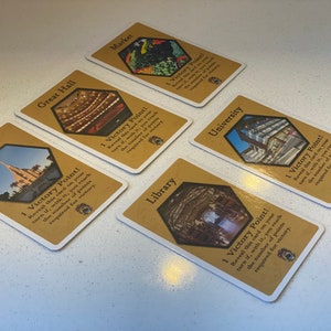 Puede incluir: Cinco cartas de juego de cartas diferentes con un fondo marr&oacute;n y una imagen hexagonal de un edificio en cada carta. Cada carta tiene el texto "&iexcl;1 punto de victoria!" e instrucciones sobre c&oacute;mo ganar el punto.
