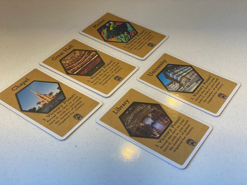 Puede incluir: Cinco cartas de juego de cartas diferentes con un fondo marr&oacute;n y una imagen hexagonal de un edificio en cada carta. Cada carta tiene el texto "&iexcl;1 punto de victoria!" e instrucciones sobre c&oacute;mo ganar el punto.