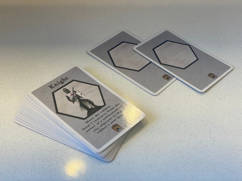 Puede incluir: Una pila de cartas de juego grises y blancas con una ilustraci&oacute;n de caballero y el texto "Knight" en la carta superior. El texto de la carta dice "Move the robber. Steal 1 resource from the owner of a settlement or city adjacent to the robber's new hex."