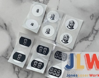 May include: A set of round stickers featuring black and white illustrations. Some stickers have a black background with white illustrations of a hand in a glove, while others have a white background with black illustrations of a person wearing a helmet. The stickers are arranged on a white surface.