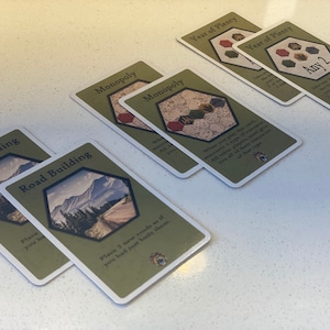 Puede incluir: Primer plano de seis cartas de juego dispuestas sobre una superficie blanca. Las cartas son verdes y blancas con im&aacute;genes de monta&ntilde;as, carreteras y una cuadr&iacute;cula hexagonal. El texto en las cartas incluye "Construcci&oacute;n de carreteras", "Monopolio" y "A&ntilde;o de abundancia".