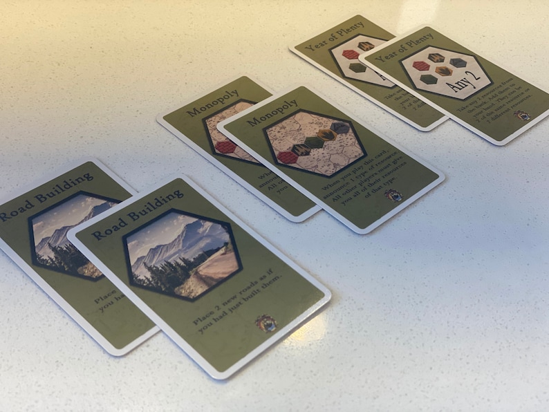 Puede incluir: Primer plano de seis cartas de juego dispuestas sobre una superficie blanca. Las cartas son verdes y blancas con im&aacute;genes de monta&ntilde;as, carreteras y una cuadr&iacute;cula hexagonal. El texto en las cartas incluye "Construcci&oacute;n de carreteras", "Monopolio" y "A&ntilde;o de abundancia".