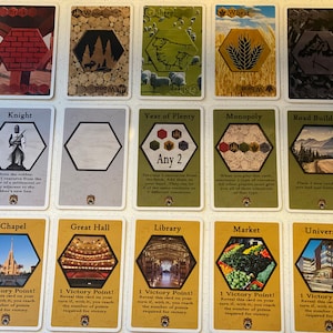 Puede incluir: Un conjunto de 12 cartas de juego Catan que representan varias cartas de recursos y desarrollo. Las cartas son amarillas, verdes y marrones con formas hexagonales e im&aacute;genes de recursos como madera, ovejas, trigo y ladrillo. Las cartas de desarrollo incluyen Caballero, A&ntilde;o de la Abundancia, Monopolio, Construcci&oacute;n de Carreteras, Capilla, Gran Sal&oacute;n, Biblioteca, Mercado y Universidad.