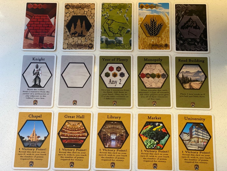 Puede incluir: Un conjunto de 12 cartas de juego Catan que representan varias cartas de recursos y desarrollo. Las cartas son amarillas, verdes y marrones con formas hexagonales e im&aacute;genes de recursos como madera, ovejas, trigo y ladrillo. Las cartas de desarrollo incluyen Caballero, A&ntilde;o de la Abundancia, Monopolio, Construcci&oacute;n de Carreteras, Capilla, Gran Sal&oacute;n, Biblioteca, Mercado y Universidad.