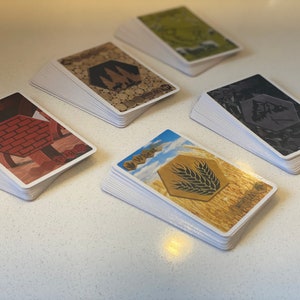 Puede incluir: Cinco pilas de cartas con fondos e im&aacute;genes de diferentes colores. Las cartas est&aacute;n etiquetadas con los nombres de diferentes recursos: Ladrillo, Madera, Mineral, Trigo y Oveja.