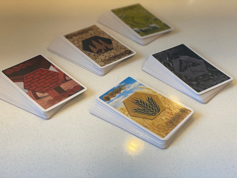 Puede incluir: Cinco pilas de cartas con fondos e im&aacute;genes de diferentes colores. Las cartas est&aacute;n etiquetadas con los nombres de diferentes recursos: Ladrillo, Madera, Mineral, Trigo y Oveja.