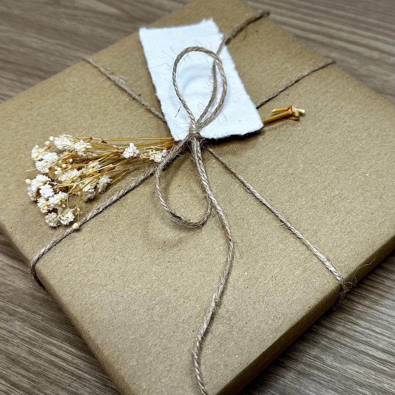 Pu&ograve; includere: Un pacco quadrato avvolto in carta marrone, legato con spago e adornato con un'etichetta di carta bianca e fiori secchi. Il pacco suggerisce un regalo o un oggetto speciale.