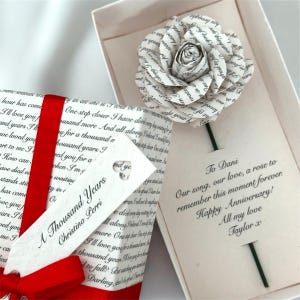 Puede incluir: Una rosa de papel hecha con páginas de libro, presentada en una caja de regalo. La caja está adornada con una cinta roja e incluye una etiqueta con el texto "A Thousand Years" de Christina Perri. También se incluye un mensaje escrito a mano.