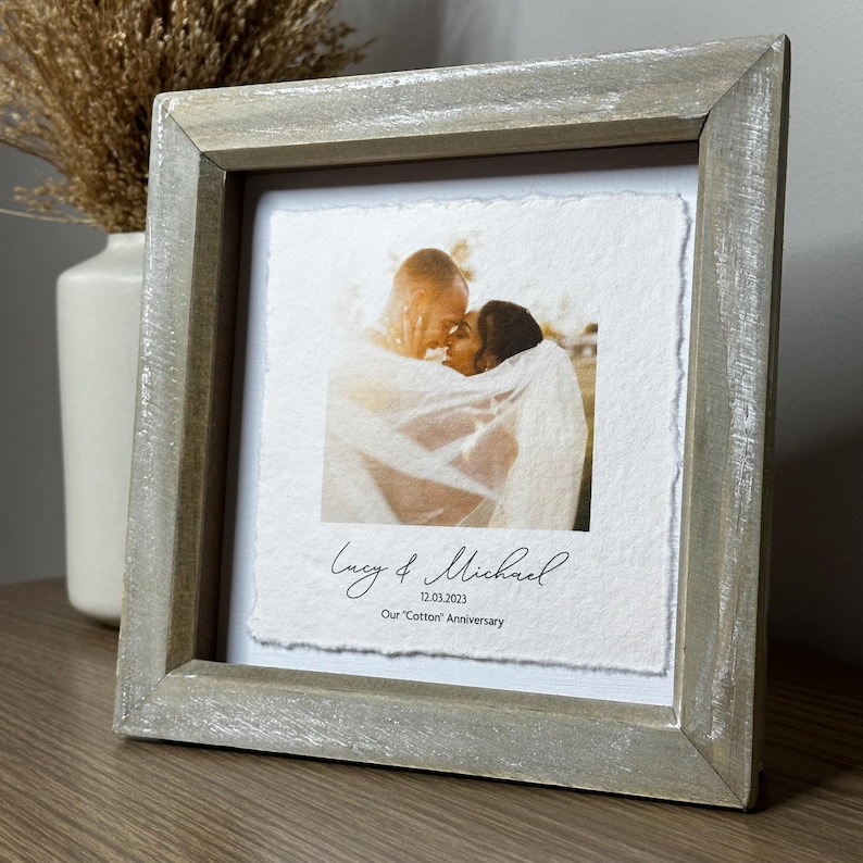 Pu&ograve; includere: Una fotografia incorniciata con una coppia che si abbraccia, con i nomi "Lucy & Michael" e la data "12.03.2023" sotto. La cornice &egrave; in legno grigio chiaro e la foto &egrave; su carta ruvida. Il testo recita "Our 'Cotton' Anniversary".