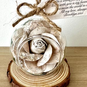 以下が含まれることがあります： 木のスライスの上に置かれた、中に紙のバラが入った透明なガラスの球体。バラは地図のページで作られています。手書きのテキストが入ったタグが紐で上部に結ばれています。テキストは「In a world full of...」と「All my love, always Brooklyn xxx」と書かれています。