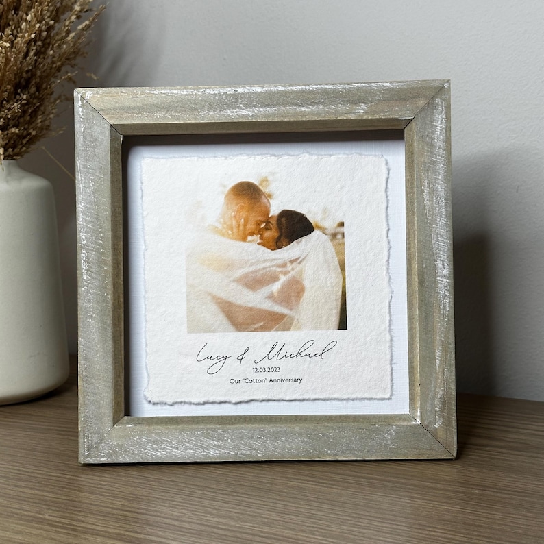 Pu&ograve; includere: Una fotografia incorniciata con una coppia che si abbraccia, su uno sfondo bianco strutturato. La cornice &egrave; in legno grigio chiaro. I nomi "Lucy & Michael" e la data "12.03.2021" sono scritti sotto la foto, con la frase "Our 'Cotton' Anniversary".