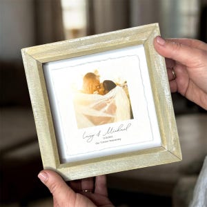 Pu&ograve; includere: Una cornice quadrata in legno chiaro che contiene una fotografia di una coppia. La foto &egrave; centrata su uno sfondo bianco con i nomi "Lucy & Michael" e la data "12.05.2023" stampati sotto. &Egrave; stampato anche il testo "Our 'Cotton' Anniversary".