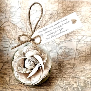 Puede incluir: Un adorno de cristal transparente lleno de pétalos de rosa de papel y un anillo plateado. El adorno está atado con hilo y una etiqueta que dice: "One chapter ends, another begins. May retirement bring joy, freedom, and new journeys. Love, Kerry & Sam xxx."