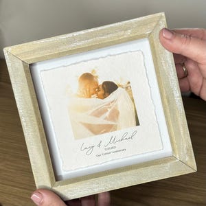 Pu&ograve; includere: Cornice quadrata in legno chiaro che contiene una fotografia di una coppia che si abbraccia. La foto &egrave; su carta strutturata e include i nomi "Lucy & Michael" e la data "12.03.2023", con il testo "Our 'Cotton' Anniversary" sotto.