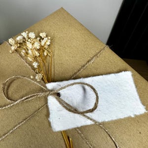 Pu&ograve; includere: Un regalo avvolto in carta marrone, legato con spago e adornato con fiori bianchi secchi e un'etichetta bianca vuota. Lo spago &egrave; annodato a fiocco attorno ai fiori e all'etichetta, creando un'estetica rustica e naturale.