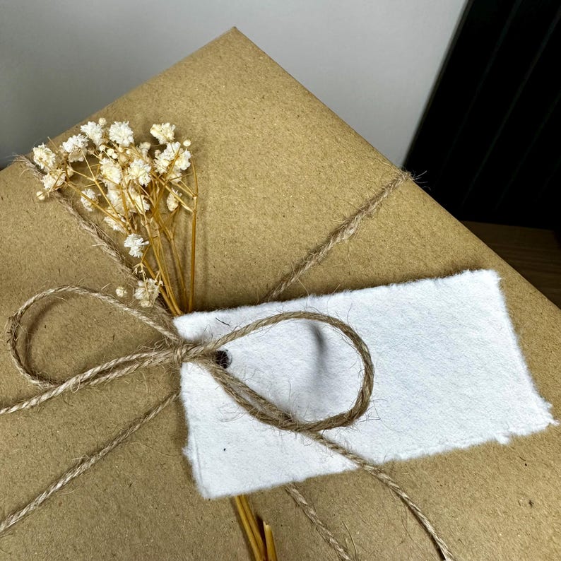 Pu&ograve; includere: Un regalo avvolto in carta marrone, legato con spago e adornato con fiori bianchi secchi e un'etichetta bianca vuota. Lo spago &egrave; annodato a fiocco attorno ai fiori e all'etichetta, creando un'estetica rustica e naturale.