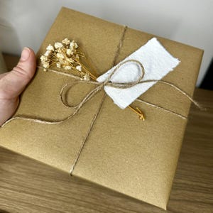 Pu&ograve; includere: Un pacco quadrato avvolto in carta marrone, legato con spago e adornato con un rametto di fiori secchi e un'etichetta di carta bianca. Il pacco &egrave; presentato su una superficie di legno.