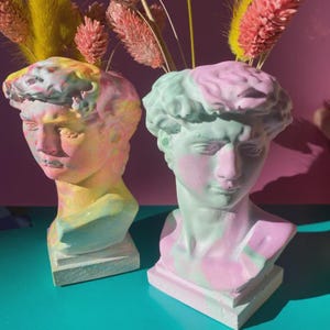 May include: Two decorative bust vases, each painted in pastel colors. One is a mix of yellow, pink, and blue, while the other is primarily mint green and pink. Both are on a square base and hold dried flowers.
