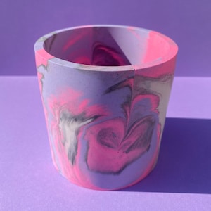 May include: A cylindrical planter with a marbled design in shades of pink, purple, and gray. The planter is made of concrete and has a smooth, polished finish.