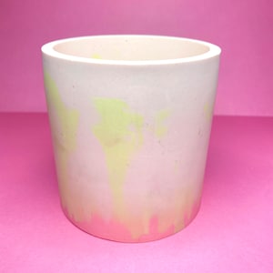 May include: A white ceramic planter with a light green and pink ombre design. The planter has a cylindrical shape and a smooth, matte finish.
