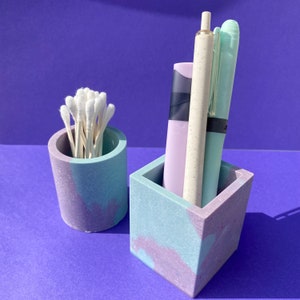 May include: Two small, decorative containers made of concrete with a marbled, light blue and purple design. One container holds cotton swabs and the other holds a pen and a highlighter.