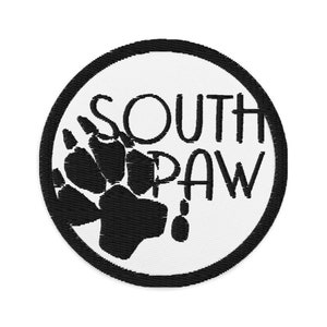Pode incluir: Etiqueta bordada em preto e branco com uma pegada de pata e o texto "SOUTH PAW" em um círculo.