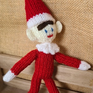 Puede incluir: Una muñeca de elfo de punto con un gorro rojo y blanco, un atuendo rojo y blanco y una cara blanca con ojos azules. El elfo está de pie sobre una superficie de madera.