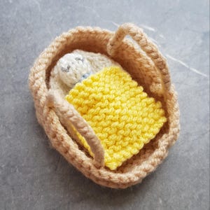 May include: A small knitted doll in a yellow blanket is lying in a beige woven basket with handles.