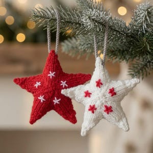 Pode incluir: Dois ornamentos em forma de estrela de cinco pontas feitos de tecido de malha. Uma estrela é vermelha com detalhes bordados em ponto cruz branco, e a outra é branca com detalhes bordados em ponto cruz vermelho. Cada estrela pende de um laço cinza.