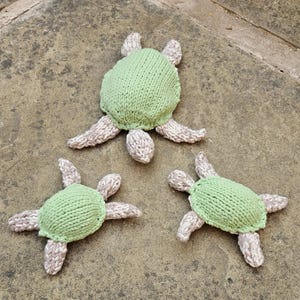 May include: Three knitted turtle toys. Each turtle has a light green shell and beige flippers and head. The toys are arranged on a textured, neutral-toned surface.