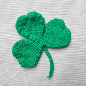 Puede incluir: Un trébol tejido de color verde brillante que presenta tres hojas en forma de corazón y un tallo delgado. La pieza está elaborada con un patrón de punto jersey visible, creando una superficie texturizada que imita la apariencia de una hoja.