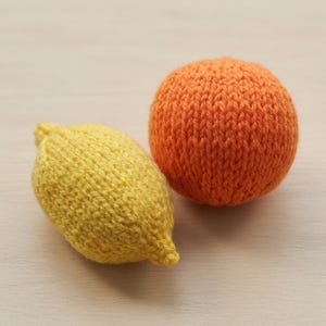 May include: A hand-knitted lemon-shaped object in bright yellow yarn and a spherical orange-colored knitted object. Both items feature a visible stockinette stitch texture and are crafted from soft yarn, displaying distinct fruit-like shapes.